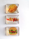 Three rectangular clear fridge magnets stacked vertically on a white background, each printed with a different food illustration: a baked potato with cheese, a grilled skewer with red peppers, and a hamburger with lettuce and tomato.