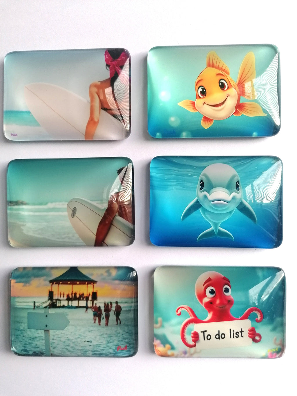 Six rectangular fridge magnets with rounded corners arranged in a 2x3 grid on a white background. Each coaster features a different colourful printed design: a surfer, a yellow fish, a dolphin, a beach pier scene, a red octopus holding a 'To do list' sign, and a turquoise underwater scene.