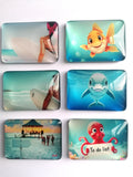 Six rectangular fridge magnets with rounded corners arranged in a 2x3 grid on a white background. Each coaster features a different colourful printed design: a surfer, a yellow fish, a dolphin, a beach pier scene, a red octopus holding a 'To do list' sign, and a turquoise underwater scene.